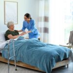 An elderly woman in a bed, receiving a glass of water from a woman in blue scrubs. To the side of the bed is a walker.