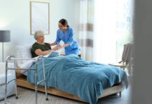 An elderly woman in a bed, receiving a glass of water from a woman in blue scrubs. To the side of the bed is a walker.