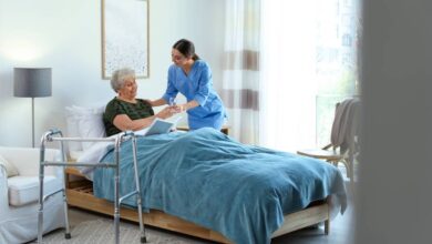 An elderly woman in a bed, receiving a glass of water from a woman in blue scrubs. To the side of the bed is a walker.