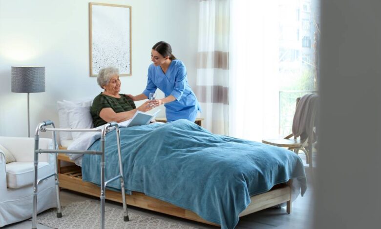 An elderly woman in a bed, receiving a glass of water from a woman in blue scrubs. To the side of the bed is a walker.