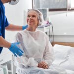 A woman in a disposable gown sitting on a surgery table and smiling up at a man standing over her with a hand on her back.