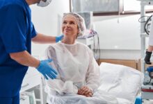 A woman in a disposable gown sitting on a surgery table and smiling up at a man standing over her with a hand on her back.