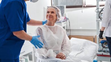 A woman in a disposable gown sitting on a surgery table and smiling up at a man standing over her with a hand on her back.