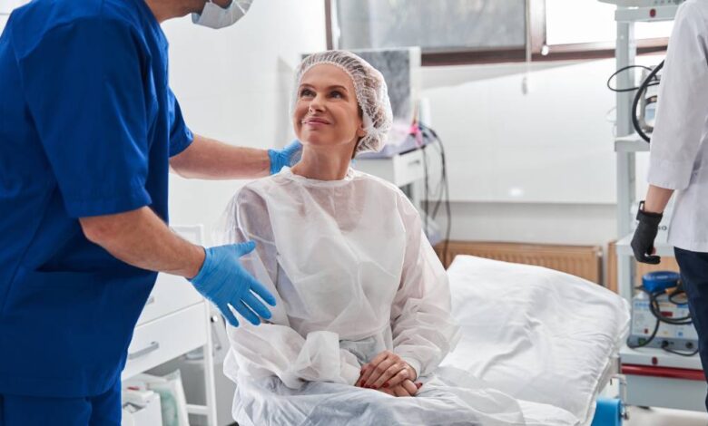 A woman in a disposable gown sitting on a surgery table and smiling up at a man standing over her with a hand on her back.