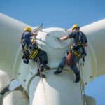 Two people hanging on the side of a wind turbine. They're both wearing harnessing gear and helmets.