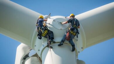 Two people hanging on the side of a wind turbine. They're both wearing harnessing gear and helmets.