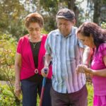 A woman in a purple shirt helps an older man walk through a wooded area. An older woman walks behind them.