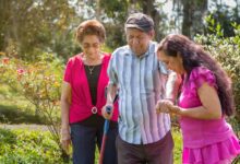 A woman in a purple shirt helps an older man walk through a wooded area. An older woman walks behind them.