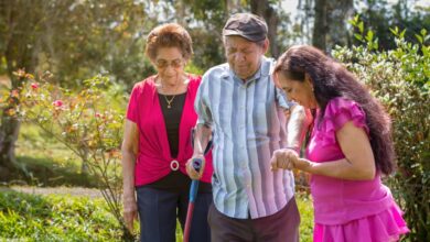 A woman in a purple shirt helps an older man walk through a wooded area. An older woman walks behind them.