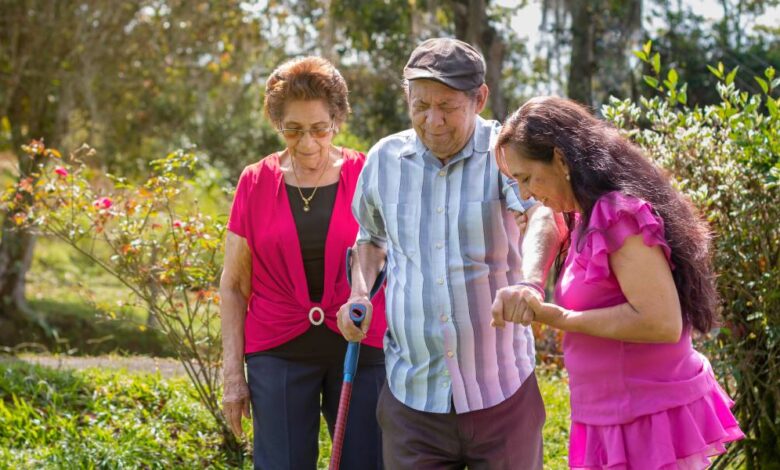 A woman in a purple shirt helps an older man walk through a wooded area. An older woman walks behind them.