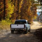 A white truck drives down a dusty dirt road surrounded by tall autumn trees on the side of the road.