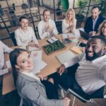 A group of employees gathered around a small conference table. They are all smiling and taking a selfie together.