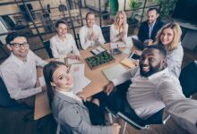 A group of employees gathered around a small conference table. They are all smiling and taking a selfie together.