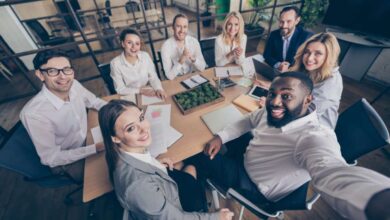 A group of employees gathered around a small conference table. They are all smiling and taking a selfie together.