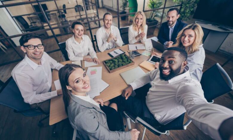 A group of employees gathered around a small conference table. They are all smiling and taking a selfie together.