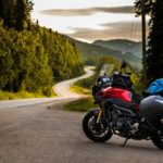 A motorcycle parked on the side of a curvy, twisty road. The sun is going down behind the tree line.