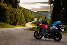 A motorcycle parked on the side of a curvy, twisty road. The sun is going down behind the tree line.