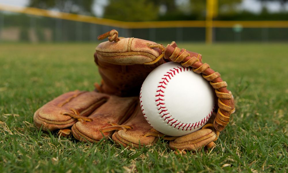 Baseball sitting inside of a baseball glove while lying in the outfield. Slightly cloudy skies with trees in the background.