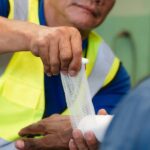 A factory worker in a yellow safety vest wrapping another worker's wrist and hand with a white bandage.