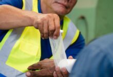 A factory worker in a yellow safety vest wrapping another worker's wrist and hand with a white bandage.