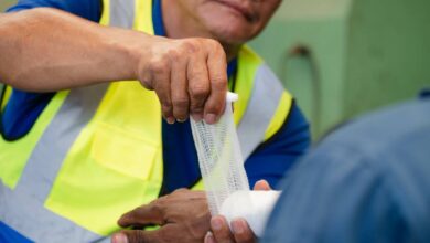 A factory worker in a yellow safety vest wrapping another worker's wrist and hand with a white bandage.