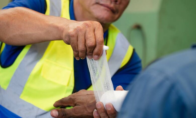 A factory worker in a yellow safety vest wrapping another worker's wrist and hand with a white bandage.