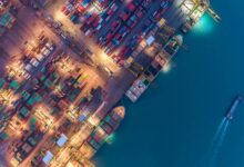 An overhead view shows a shipping cargo port. Large cranes are moving containers onto large cargo ships, and lights are on.