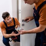 Two professional locksmiths working on a residential interior door handle. They're in orange shirts and coveralls.