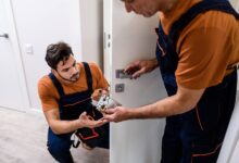 Two professional locksmiths working on a residential interior door handle. They're in orange shirts and coveralls.