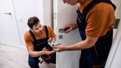 Two professional locksmiths working on a residential interior door handle. They're in orange shirts and coveralls.