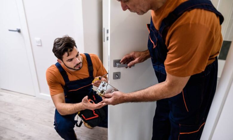 Two professional locksmiths working on a residential interior door handle. They're in orange shirts and coveralls.