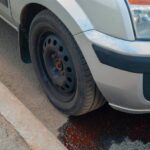 A car parked next to a sidewalk. There's red fluid leaking from underneath the car, pooling by the front tire.