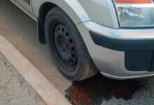 A car parked next to a sidewalk. There's red fluid leaking from underneath the car, pooling by the front tire.