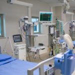 A row of hospital beds with small windows above them surrounded by different types of medical equipment.