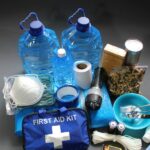 A pile of essentials including gallons of water, a mask, rations, a flashlight, rope, matches, and a first aid kit.
