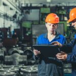 Two workers wearing helmets and blue uniforms inside a manufacturing room holding and looking at a tablet.