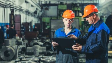 Two workers wearing helmets and blue uniforms inside a manufacturing room holding and looking at a tablet.