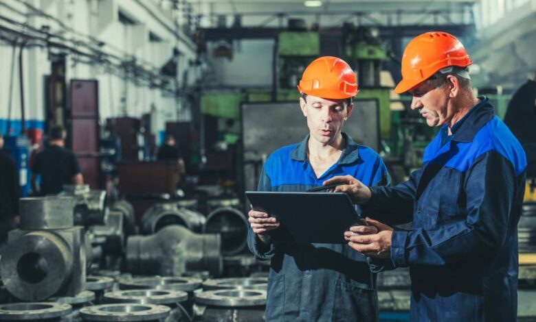 Two workers wearing helmets and blue uniforms inside a manufacturing room holding and looking at a tablet.