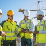 A group of four construction professionals discussing a plan at the job site. An industrial crane stands in the distance.