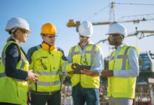 A group of four construction professionals discussing a plan at the job site. An industrial crane stands in the distance.