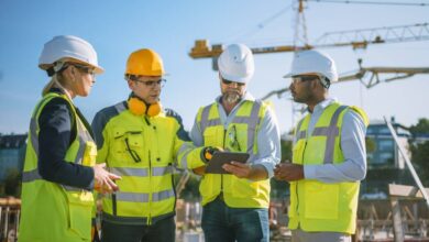 A group of four construction professionals discussing a plan at the job site. An industrial crane stands in the distance.