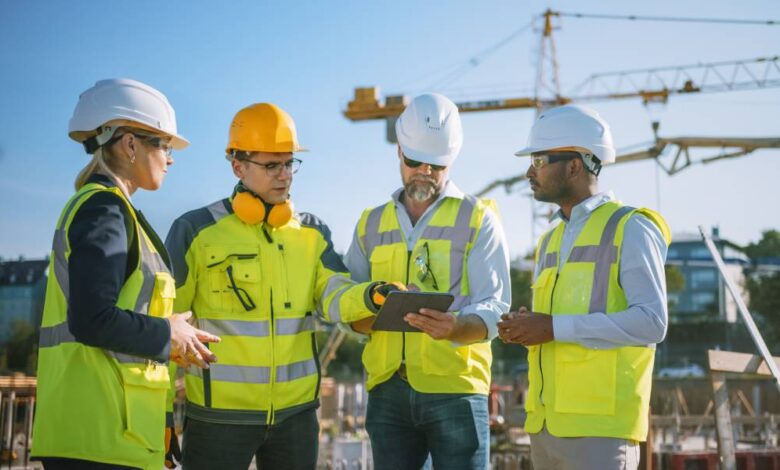 A group of four construction professionals discussing a plan at the job site. An industrial crane stands in the distance.