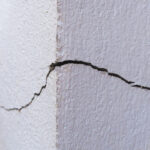 Close-up of a corner of a building that features a crack on both sides. The corner is white and has plants alongside it.