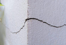 Close-up of a corner of a building that features a crack on both sides. The corner is white and has plants alongside it.