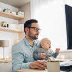 A man holds a baby in his lap while working on a desktop computer in a home office space.