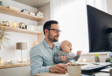 A man holds a baby in his lap while working on a desktop computer in a home office space.