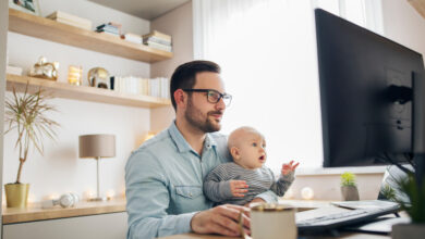 A man holds a baby in his lap while working on a desktop computer in a home office space.