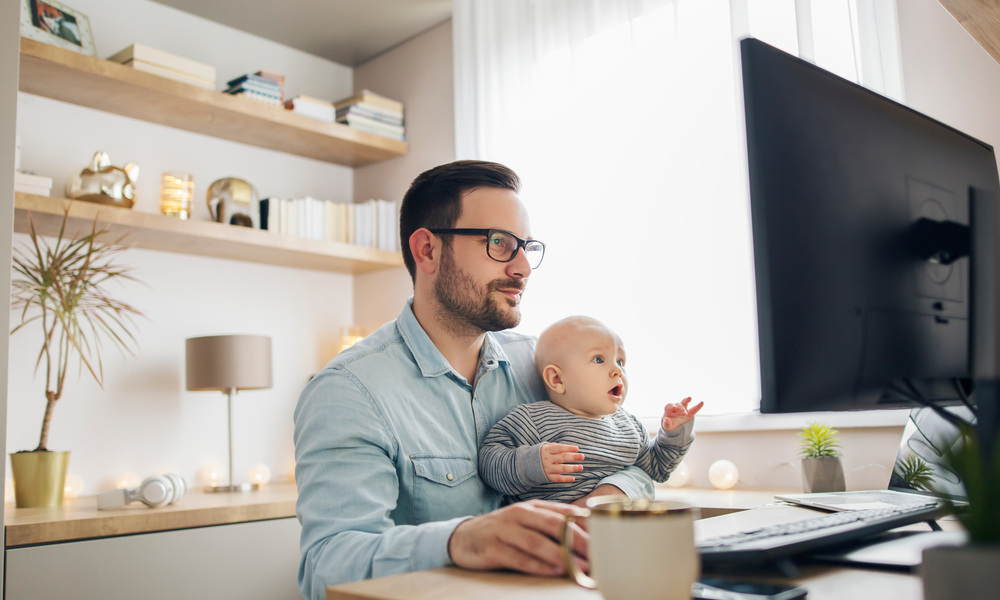 A man holds a baby in his lap while working on a desktop computer in a home office space.
