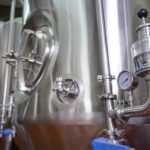 Stainless steel industrial holding tanks housed inside a warehouse. They have pressure gauges and pipework on the bottom.
