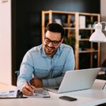 A young professional working at his home office—he productively writes on a pad of paper while on his laptop.